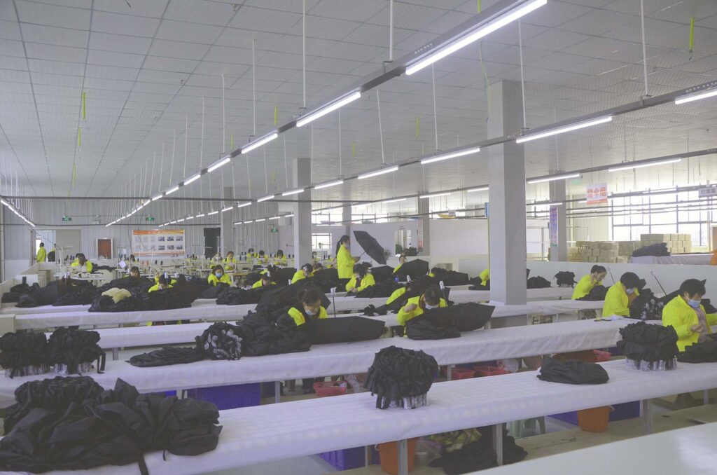 Workers in yellow jackets working on black umbrellas at long white tables in a large workshop