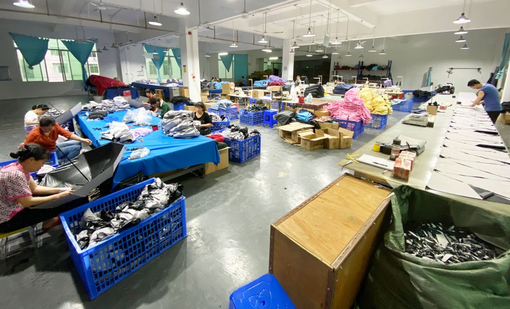 Workshop floor where workers handle umbrella canopies at blue tables beside crates of umbrella parts